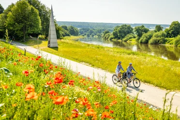 Hoexter-Weserradweg-Stadt Hoexter-D. Ketz-2023-312.jpg Zwei Radfahrer fahren auf einem Weg entlang des Flusses, umgeben von blühenden Mohnblumen und grünen Wiesen.