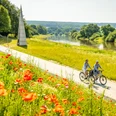 Hoexter-Weserradweg-Stadt Hoexter-D. Ketz-2023-312.jpg Zwei Radfahrer fahren auf einem Weg entlang des Flusses, umgeben von blühenden Mohnblumen und grünen Wiesen.