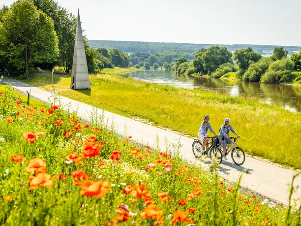 Hoexter-Weserradweg-Stadt Hoexter-D. Ketz-2023-312.jpg Zwei Radfahrer fahren auf einem Weg entlang des Flusses, umgeben von blühenden Mohnblumen und grünen Wiesen.
