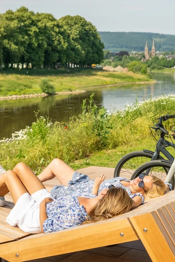 Hoexter-Weserradweg-Stadt Hoexter-D. Ketz-2023-300.jpg Zwei Radfahrerinnen machen Pause auf Holzliege auf der Weserscholle