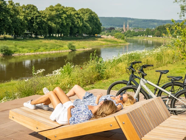 Hoexter-Weserradweg-Stadt Hoexter-D. Ketz-2023-300.jpg Zwei Radfahrerinnen machen Pause auf Holzliege auf der Weserscholle