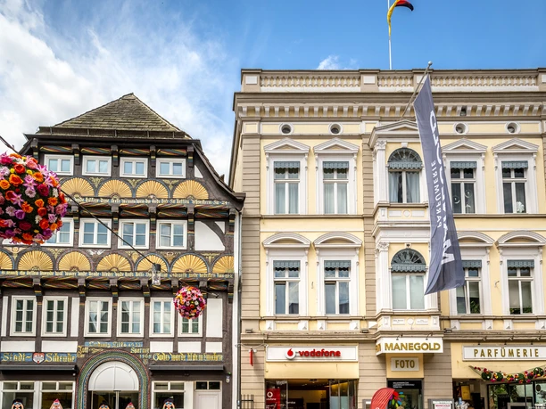 Hoexter-Stadtfuehrung-Stadt Hoexter-D. Ketz-2023-128.jpg Zwei historische Fassaden in Höxter, verziert mit bunten Blumenampeln und einer Flagge im Hintergrund.