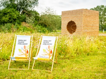 Hoexter-Landesgartenschaugelände Archeologiepark-Stadt Hoexter-D. Ketz-2023-371.jpg Holzkubus aus gekreuzt geschichteten Holzlatten mit rundem Loch an einer Seite und Stühlen im Vordergrund auf einer Wiese