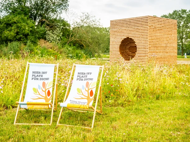 Hoexter-Landesgartenschaugelände Archeologiepark-Stadt Hoexter-D. Ketz-2023-371.jpg Holzkubus aus gekreuzt geschichteten Holzlatten mit rundem Loch an einer Seite und Stühlen im Vordergrund auf einer Wiese