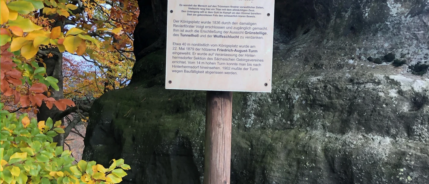 Bord op Königsplatz bij Hinterhermsdorf Houten bord met tekst voor een grote rots, omringd door herfstkleurige bomen. De sfeer is rustig en natuurlijk.