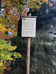 Schild am Königsplatz bei Hinterhermsdorf Holzschild mit Text vor einem großen Felsen, umgeben von herbstlich gefärbten Bäumen. Die Atmosphäre ist ruhig und natürlich.