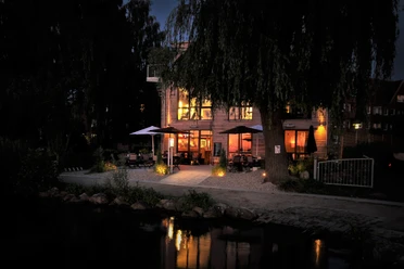 Café Köbke am Abend - Blick vom See.jpg Blick vom See auf beleuchtetes Haus mit Sitzecken und offenen Schirmen davor.