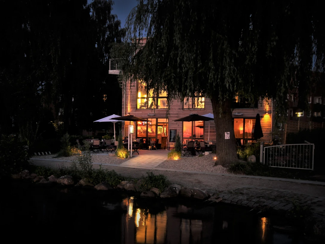 Café Köbke am Abend - Blick vom See.jpg Blick vom See auf beleuchtetes Haus mit Sitzecken und offenen Schirmen davor.
