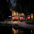 Café Köbke am Abend - Blick vom See.jpg Blick vom See auf beleuchtetes Haus mit Sitzecken und offenen Schirmen davor.