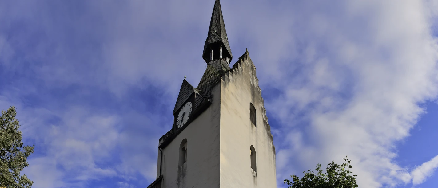 Historische Kirche mit Spitzdach und Uhrturm, umgeben von Grün, unter einem bewölkten Himmel.