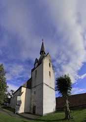 GEB1 015.jpg Historische Kirche mit Spitzdach und Uhrturm, umgeben von Grün, unter einem bewölkten Himmel.