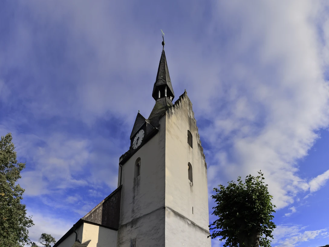 GEB1 015.jpg Historische Kirche mit Spitzdach und Uhrturm, umgeben von Grün, unter einem bewölkten Himmel.