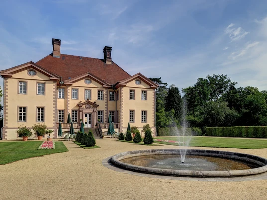 Das Schieder Schloss mit symmetrischem Park und zentralem Springbrunnen vor einem klaren, blauen Himmel.