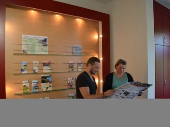 Angebot_&_Service.JPG Zwei Personen stehen in einem hellen Raum mit Broschürenwand und betrachten interessiert eine große Karte.