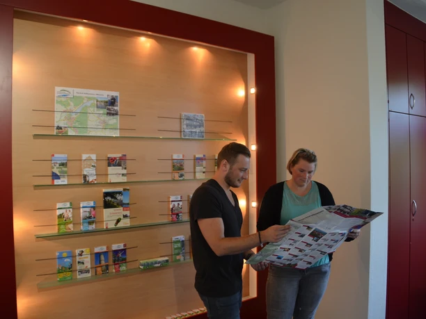 Zwei Personen betrachten in einem Informationszentrum Karten und Prospekte an einer beleuchteten Wand.