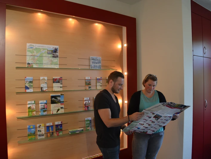 Zwei Personen betrachten in einem Informationszentrum Karten und Prospekte an einer beleuchteten Wand.