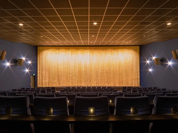 meppen_kino1_1-1280x720.jpg Kinosaal mit blauen Sitzen, goldfarbenem Vorhang und warm beleuchteten Wandleuchten