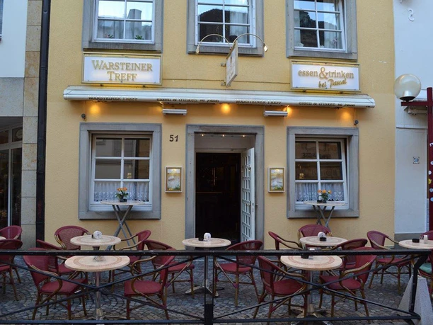 Warsteiner Treff Warsteiner Treff mit gelber Fassade, bequemen roten Stühlen und runden Tischen im Außenbereich.