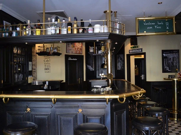 Warsteiner Treff Eine Bar mit schwarzer Holzeinrichtung und goldenen Handläufen, umliegende Barhocker und Flaschen darüber.