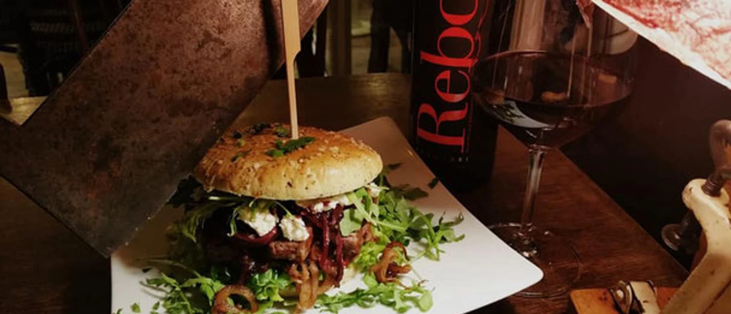 Zauber von OS Ein Gourmet-Burger liegt auf einem Teller mit Rucola, daneben ein Glas Rotwein und eine Flasche.