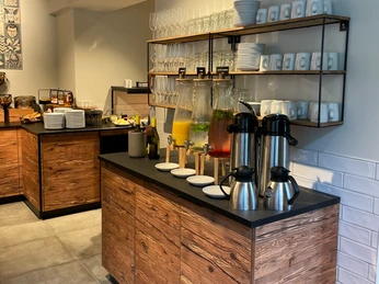Selbstbedienungsbuffet mit Getränkespendern, Kaffee, Geschirrregal und Frühstücksauswahl in modernem Ambiente.