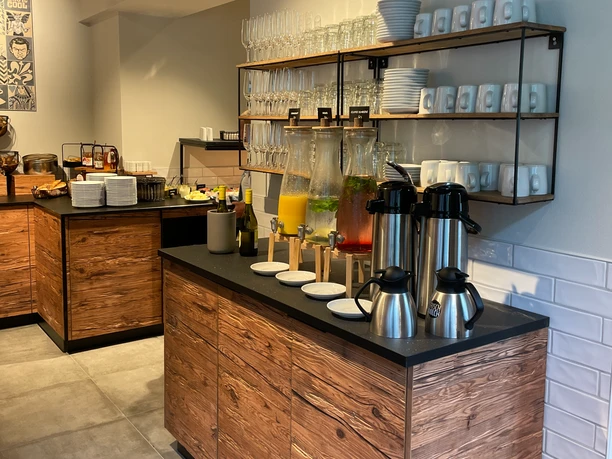 Selbstbedienungsbuffet mit Getränkespendern, Kaffee, Geschirrregal und Frühstücksauswahl in modernem Ambiente.