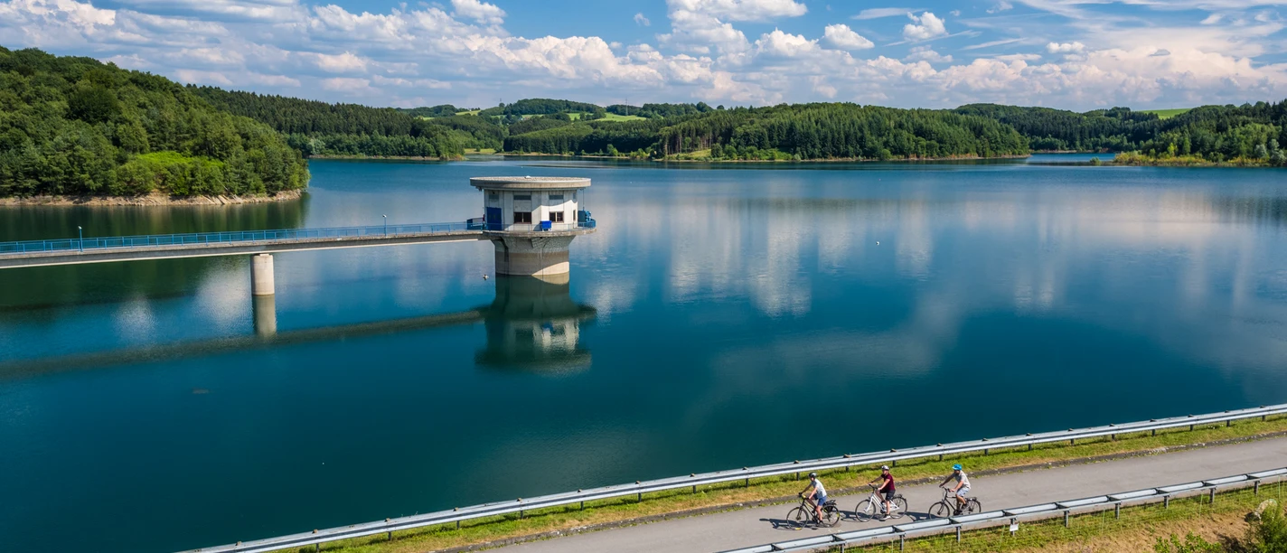 Große Dhünntalsperre Radsportler am Ufer der Talsperre Wermelskirchen, umgeben von grünen Hügeln und blauem Himmel.