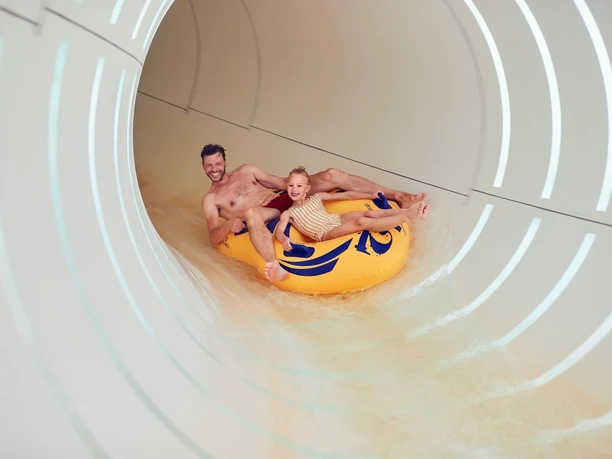 BS154909 Ein lachender Mann und ein fröhliches Kind rutschen gemeinsam in einem gelben Schlauchboot durch eine Wasserrutsche.