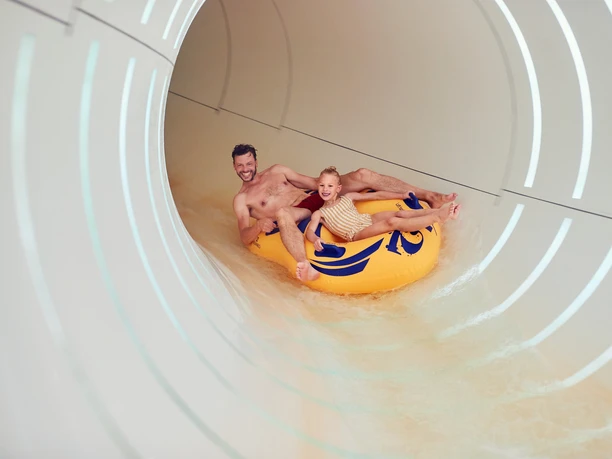 Center-parcs-bispinger-heide-8_BS154909-1.jpg Ein lachender Mann und ein fröhliches Kind rutschen gemeinsam in einem gelben Schlauchboot durch eine Wasserrutsche.