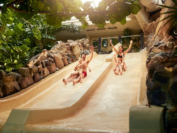 BS154922 Eine Familie rutscht lachend auf einer breiten Wasserrutsche herab, umgeben von tropischen Pflanzen und Steinen.