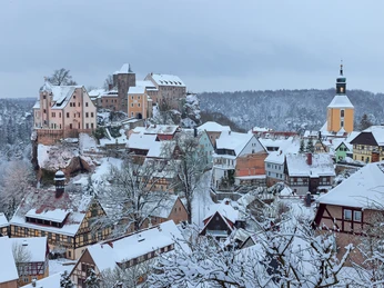 Ausblick auf Hohnstein im Winter Verschneite Altstadt mit Fachwerkhäusern und einer Burg auf einem Hügel; im Hintergrund ein Kirchturm, winterliche Atmosphäre.