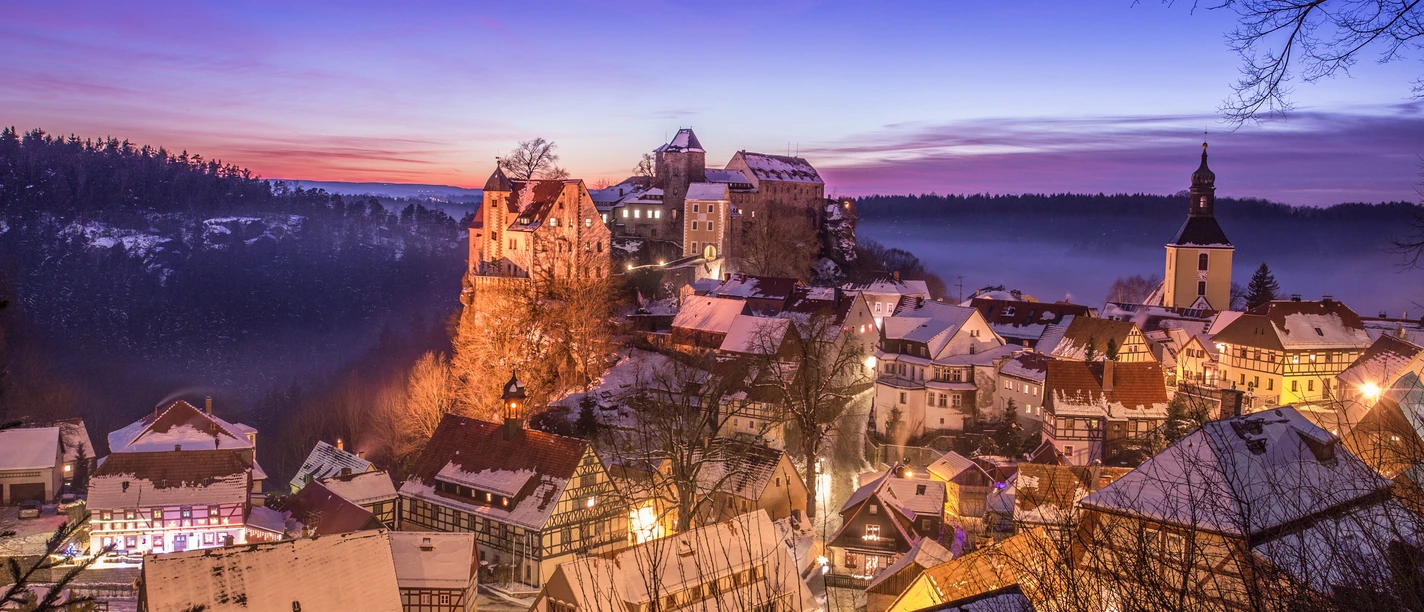 Verschneite Stadt bei Sonnenuntergang mit beleuchteten Fachwerkhäusern, einer Kirche und einer Burg auf einem Hügel, umgeben von Wald.