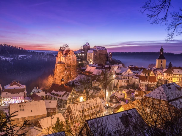 Ausblick auf Hohnstein im Winter Verschneite Stadt bei Sonnenuntergang mit beleuchteten Fachwerkhäusern, einer Kirche und einer Burg auf einem Hügel, umgeben von Wald.