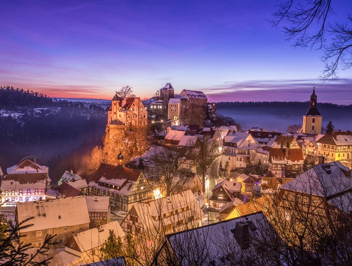 Ausblick auf Hohnstein im Winter Verschneite Stadt bei Sonnenuntergang mit beleuchteten Fachwerkhäusern, einer Kirche und einer Burg auf einem Hügel, umgeben von Wald.