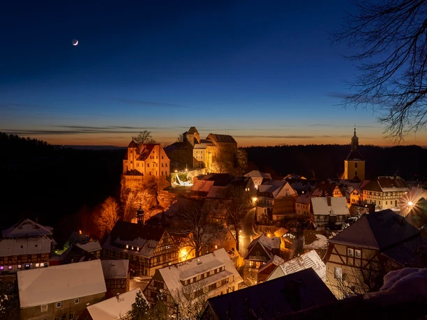 Ausblick auf Hohnstein im Winter Verschneite Stadt bei Dämmerung mit beleuchteten Fachwerkhäusern, einer Burg auf einem Hügel und einem schmalen Mond am klaren Himmel.