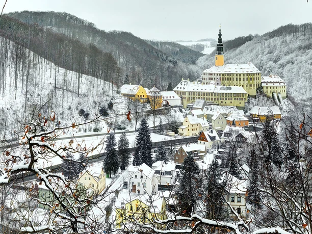 Snow-covered cityscape with pastel-colored houses and a yellow church on a hill, surrounded by snow-covered forests.