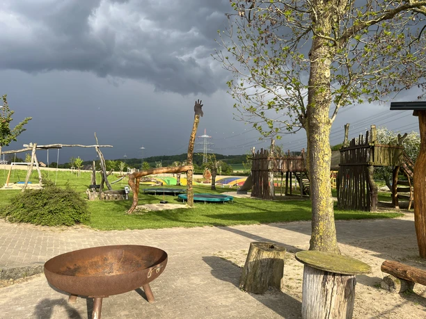 IMG_1843.JPG Ein rustikaler Spielplatz mit Holzelementen und einem Feuerkorb vor grauem, wolkenverhangenem Himmel.