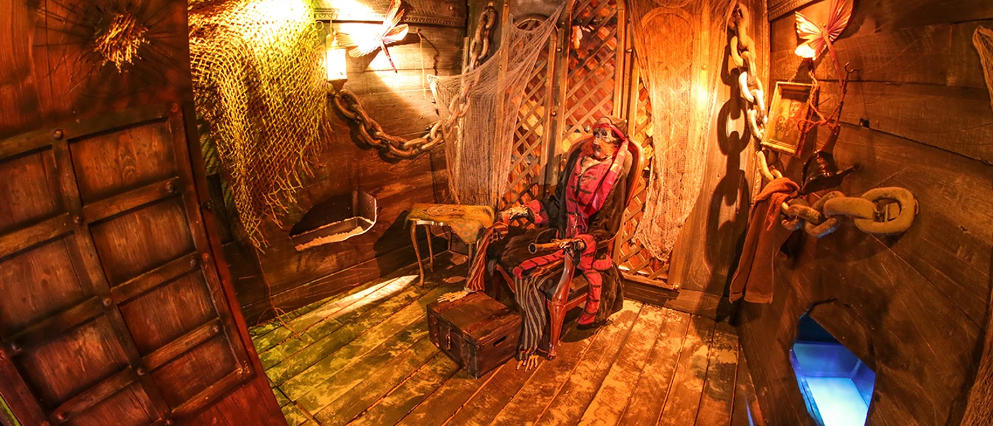 Escape Room Osnabrück.jpg Ein Piratenszenario in einem hölzernen Escape Room mit stimmungsvoller Beleuchtung und Requisiten.