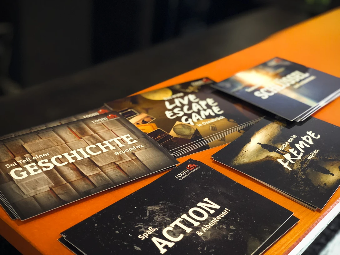 Room Fox Osnabrück.jpg Werbekarten von raumbezogenen Abenteuerspielen auf einem orangefarbenen Tisch arrangiert.