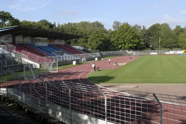 Stadion.tif Sportstadion mit Tartanbahn, Tribüne mit Sitzplätzen und angrenzendem grünen Spielfeld, umgeben von Bäumen.
