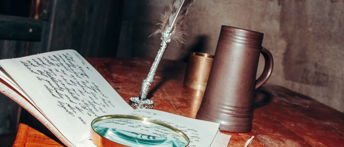 Eine Lupe, ein offenes handgeschriebenes Notizbuch, eine Feder und ein Krug auf einem Holztisch.