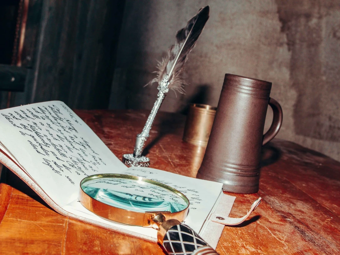 Mind Hunters Osnabrück.jpg Eine Lupe, ein offenes handgeschriebenes Notizbuch, eine Feder und ein Krug auf einem Holztisch.