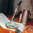 Mind Hunters Osnabrück.jpg Eine Lupe, ein offenes handgeschriebenes Notizbuch, eine Feder und ein Krug auf einem Holztisch.