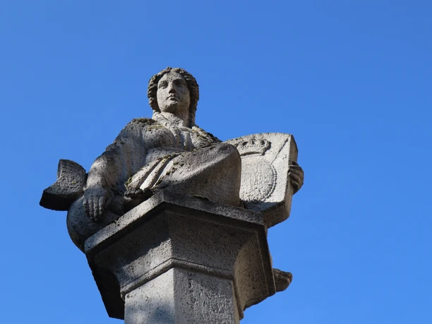 Steinfigur auf einem Brunnen sitzt hoch auf einem Sockel vor klarem, wolkenlosen Himmel.