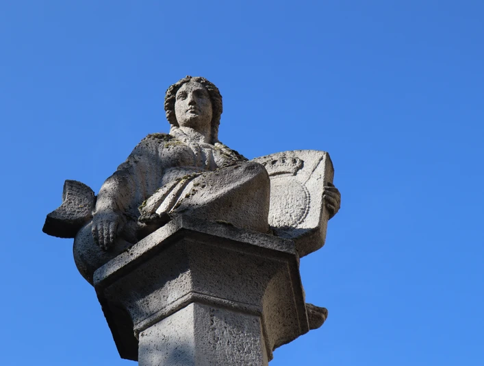 Steinfigur auf einem Brunnen sitzt hoch auf einem Sockel vor klarem, wolkenlosen Himmel.