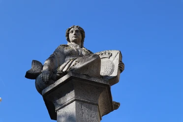 Abteibrunnen (9).JPG Steinfigur auf einem Brunnen sitzt hoch auf einem Sockel vor klarem, wolkenlosen Himmel.
