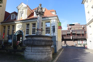 Steinbrunnen mit historischer Büste auf dem Sockel, umgeben von Architektur des 20. Jahrhunderts.