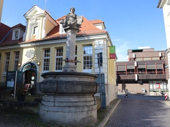 Steinbrunnen mit historischer Büste auf dem Sockel, umgeben von Architektur des 20. Jahrhunderts.