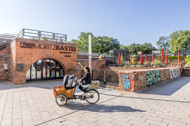 Moritzbastei - Gastronomien in Leipzig Bild vom oberen Eingang zur Moritzbastei am Kurt-Masur-Platz in Leipzig, in der es ein Café und Restaurant gibt und in dem viele Veranstaltungen stattfinden, eine Fahrradfahrerin fährt durch das Bild