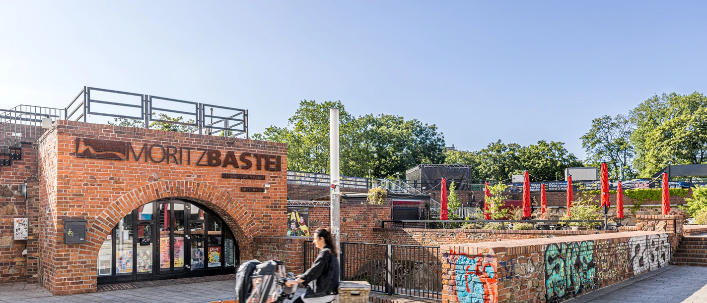 Moritzbastei - Gastronomien in Leipzig Bild vom oberen Eingang zur Moritzbastei am Kurt-Masur-Platz in Leipzig, in der es ein Café und Restaurant gibt und in dem viele Veranstaltungen stattfinden, eine Fahrradfahrerin fährt durch das Bild